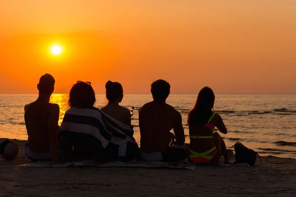 Siluetas una gente joven sentada en una playa mirando el atardecer —  Fotos de Stock