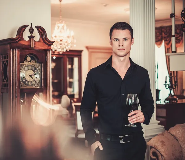 Hombre guapo joven bien vestido en el interior de la casa de lujo con copa de vino tinto — Foto de Stock