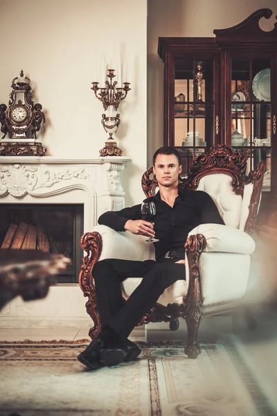Bello uomo ben vestito seduto con vino rosso in interni casa di lusso — Foto Stock