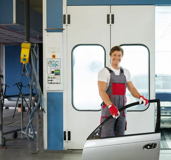 Fröhlicher Servicemann mit Autotür in der Nähe einer Lackiererei in einer Karosseriewerkstatt — Stockfoto
