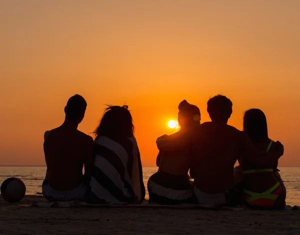 Siluetas una gente joven sentada en una playa mirando el atardecer — Foto de Stock