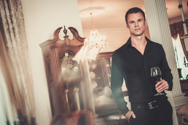 Beau jeune homme bien habillé dans l'intérieur de la maison de luxe avec un verre de vin rouge — Photo