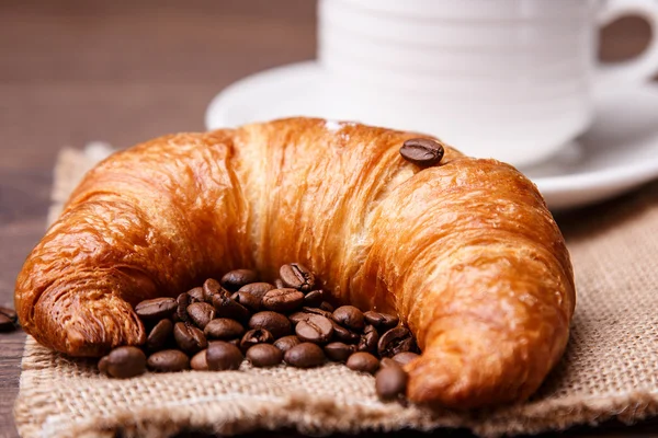 Croissant y granos de café con una taza en el fondo — Foto de Stock