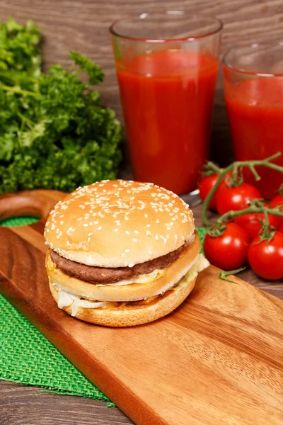 Hambúrguer saboroso em uma placa de corte e suco de tomate — Fotografia de Stock