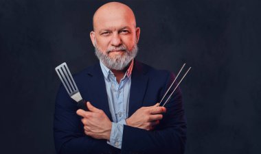 Mutfak spatulası ve maşası olan yaşlı bir adam karanlık arka plana karşı.