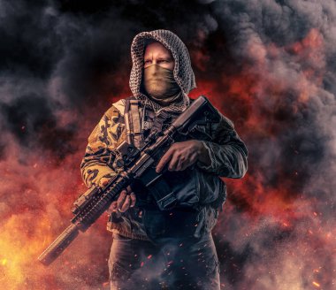 Kapüşonlu modern asker tüfeği ateşe karşı tutuyor.