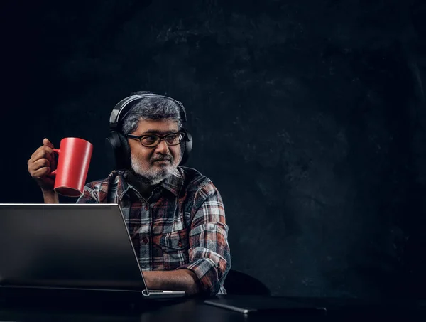 Hombre envejecido con portátil sosteniendo taza de café sobre fondo oscuro — Foto de Stock