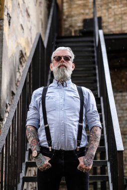 Şehirde güneş gözlüklü, şık yaşlı bir adam poz veriyor.