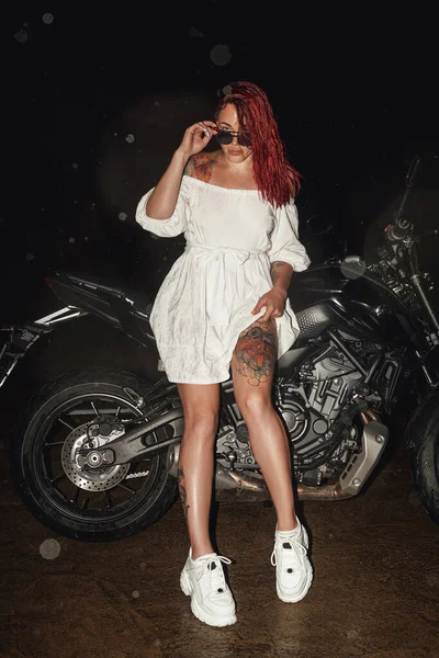 Feminino motociclista com motocicleta preta posando ao ar livre à noite — Fotografia de Stock