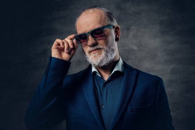 Sakallı, güneş gözlüklü yaşlı adam karanlık arka planda poz veriyor.