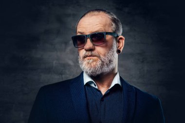 Sakallı, güneş gözlüklü yaşlı adam karanlık arka planda poz veriyor.