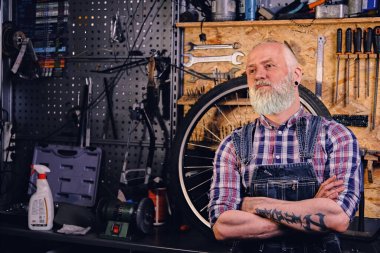 Dövmeli yaşlı bir zanaatkar atölyede çapraz kollarla diğer tarafa bakıyor.