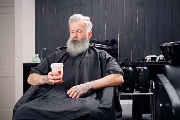 Bonito hipster velho sentado em poltrona com café na barbearia — Fotografia de Stock