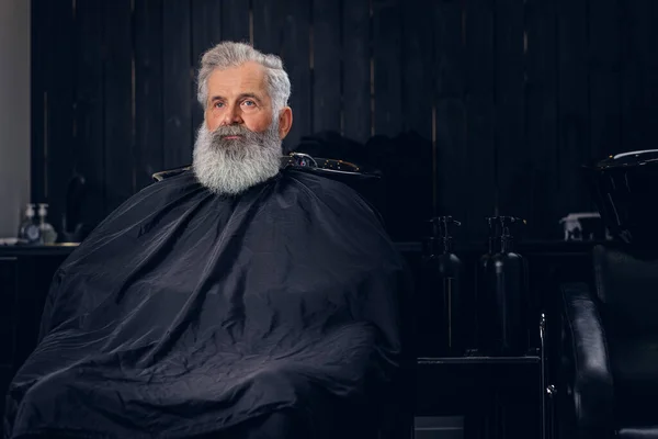 Velho hipster com peignoir sentado em poltrona na barbearia — Fotografia de Stock