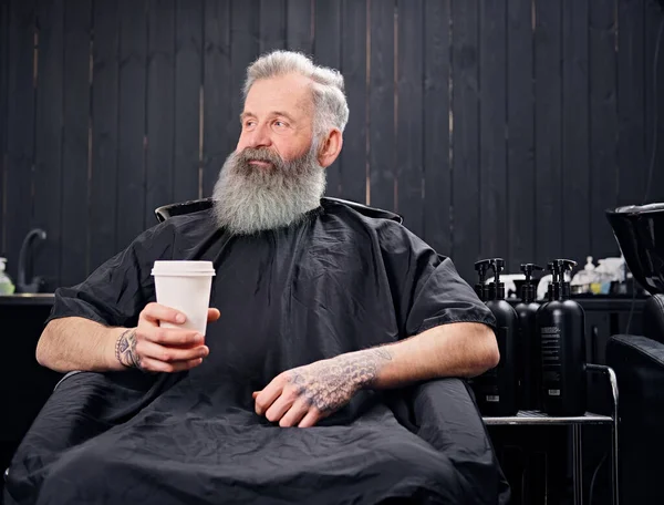 Bonito hipster velho sentado em poltrona com café na barbearia — Fotografia de Stock