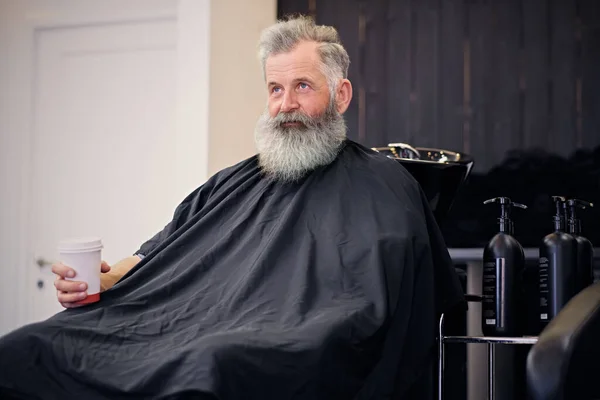 Homem elegante com longa barba e capa sentado em poltrona — Fotografia de Stock