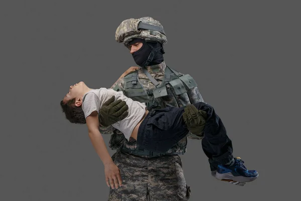 Bátor katona hazafi, sebesült gyereket cipel szürke háttérrel. — Stock Fotó