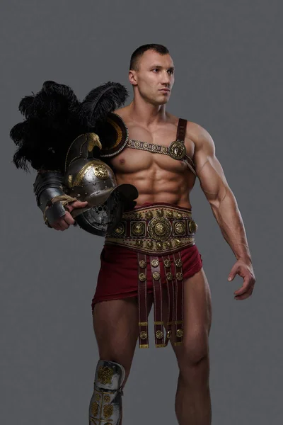 Gladiador com construção muscular segurando capacete contra fundo cinza — Fotografia de Stock