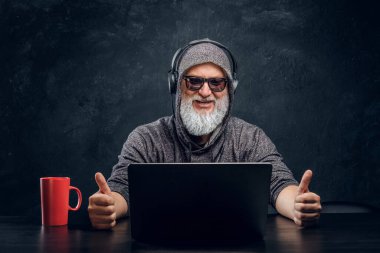 Laptop kullanan kapüşonlu ve kulaklıklı neşeli yaşlı adam.