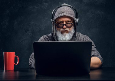 Kapüşonlu ve kulaklıklı düzenbaz yaşlı adam dizüstü bilgisayar kullanıyor.