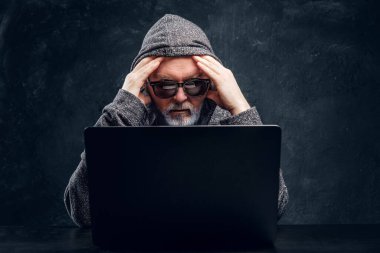 Frustrated old man hacker with laptop against dark background