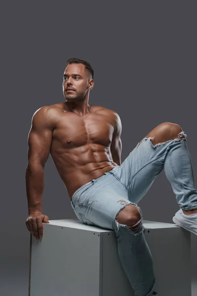 Naked bodybuilder posing sitting on box against grey background — Stock Fotó