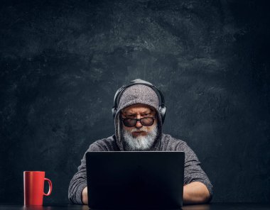 Kapüşonlu ve kulaklıklı düzenbaz yaşlı adam dizüstü bilgisayar kullanıyor.