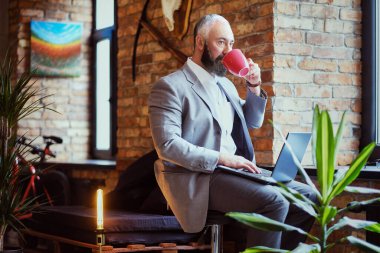 Laptoplu yaşlı adam ofisin içinde kahve fincanı içiyor.