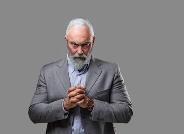 Gri takım elbiseli sakallı yaşlı adam kameraya bakıyor.