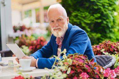 Mavi takım elbiseli, tabletli zarif yaşlı bir adam açık havada, masada oturuyor..