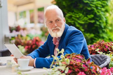 Mavi takım elbiseli, tabletli zarif yaşlı bir adam açık havada, masada oturuyor..