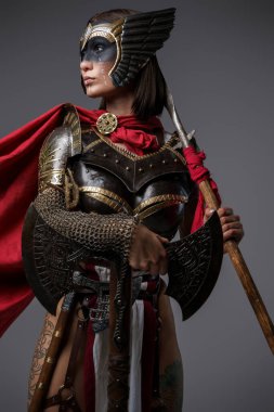 Zırhlı ve kırmızı pelerinli vahşi Amazon 'un gri arka planda balta ve mızrak tutuşu..