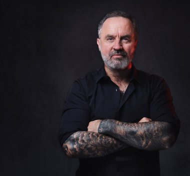 Dövmeli yaşlı hipster 'ın karanlık arka planda çapraz kollarla kameraya bakması..