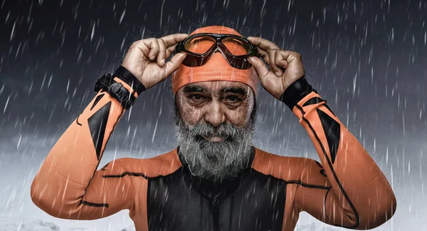 Portrait of active senior man dressed in orange swimsuit and eyewear against dark background with rain.