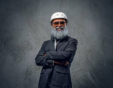 Uzun sakallı, ceketli ve kasklı, gri arka planda şık yaşlı bir adam..
