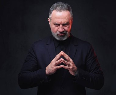 Siyah giyinmiş, siyah arka planda poz veren dalgın kıdemli bir adamın fotoğrafı..