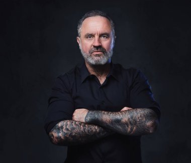 Dövmeli yaşlı hipster 'ın karanlık arka planda çapraz kollarla kameraya bakması..