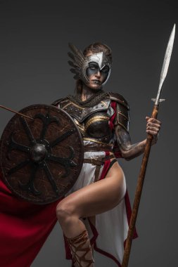 Shot of female warrior from past with shield and spear dressed in armor and red cape looking at camera.
