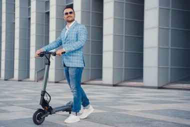 Joyful businessman with scooter dressed in blue suit and sunglasses against city building.