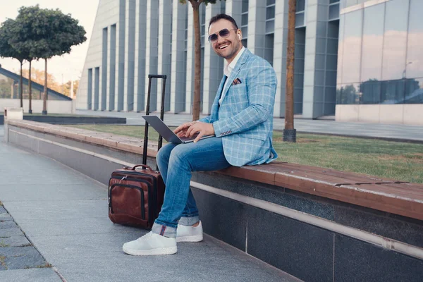 Shot of joyful businessman with suitcase and laptop looking at camera outdoors in city.