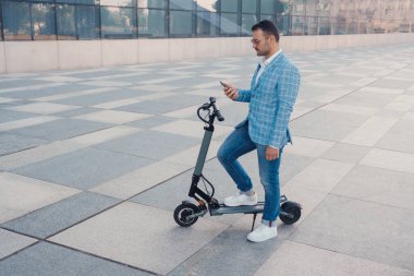 Portrait of businessman dressed in blue suit with smartphone and scooter outdoors in city.