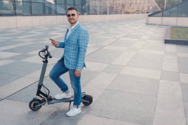 Portrait of businessman dressed in blue suit with smartphone and scooter outdoors in city.