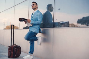 Shot of businessman with newspaper dressed in stylish blue suit leaning against city building.