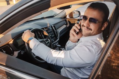 Shot of joyful businessman with sunglasses and mobile phone sitting inside expensive car.