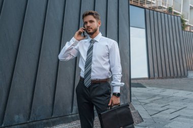 Shot of professional executive man with bag talking on phone around city buildings in daytime.