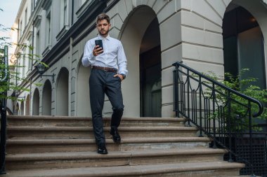 Shot of businesman dressed in modern formal attire walking in city with smartphone.