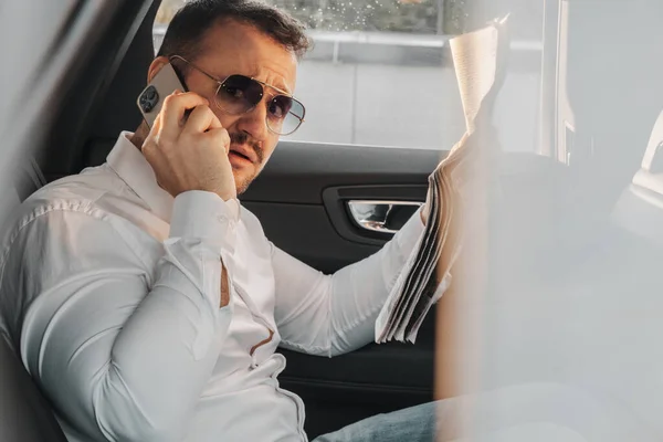 Shot of businessman with sunglasses and newspaper talking on phone sitting inside his car.