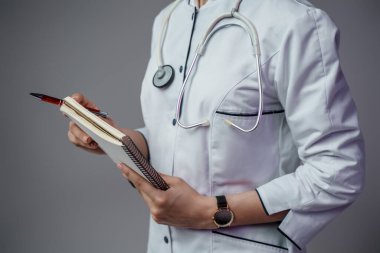Studio shot of female doctor specialist with notebook dressed in labcoat against gray background.