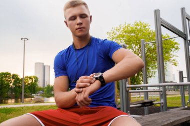 Shot of blond haired athlete dressed in sportswear setting timer on his watch outdoors in city.