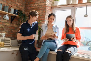 Portrait of group of three friends reading book and using tablet around window at home.
